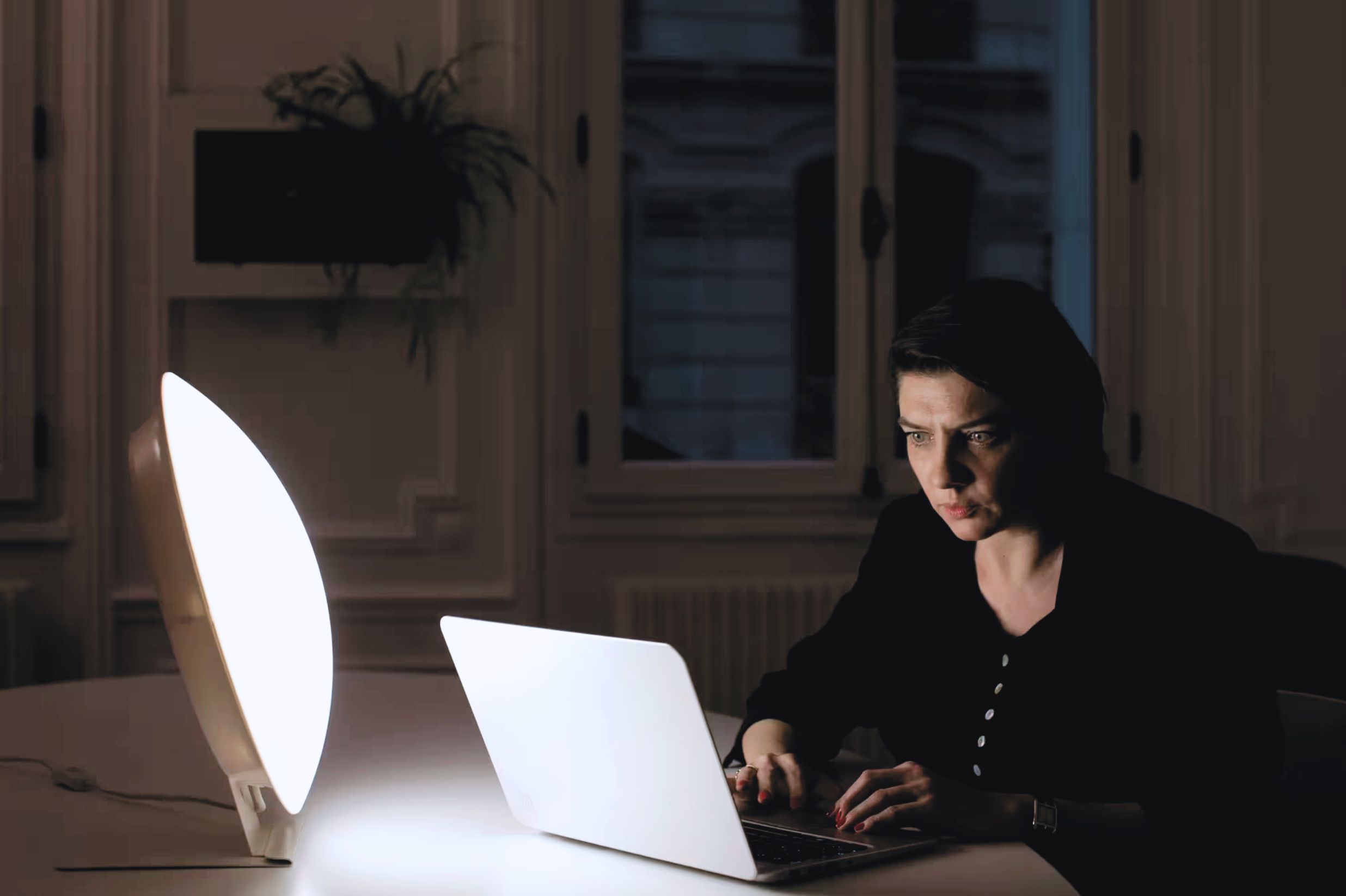 A young woman sitting at a desk looking at her laptop in a dark room with a light therapy lamp on