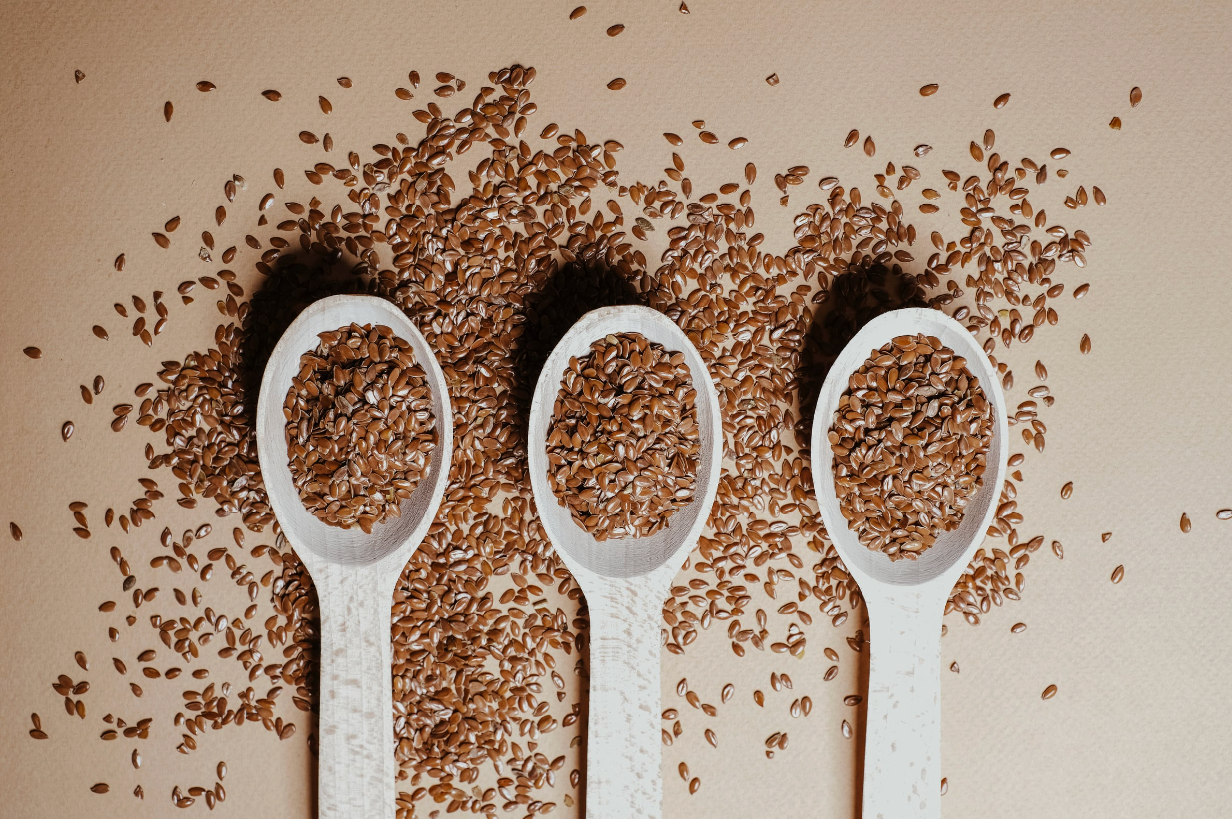 Three white plastic spoons on a beige background with flaxseeds everywhere