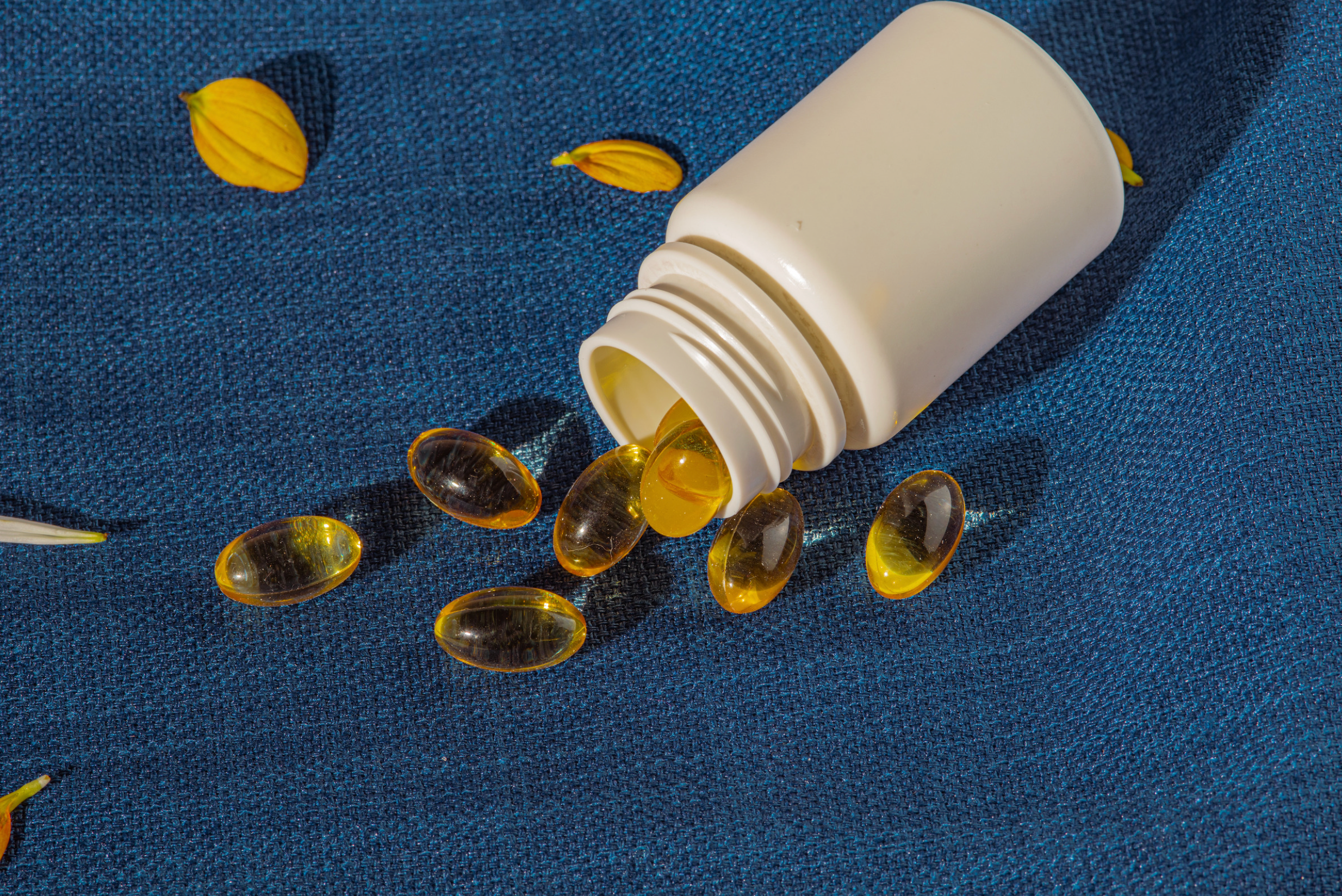 A while supplement bottle on it's side with spilled softgels, against a blue fabric background