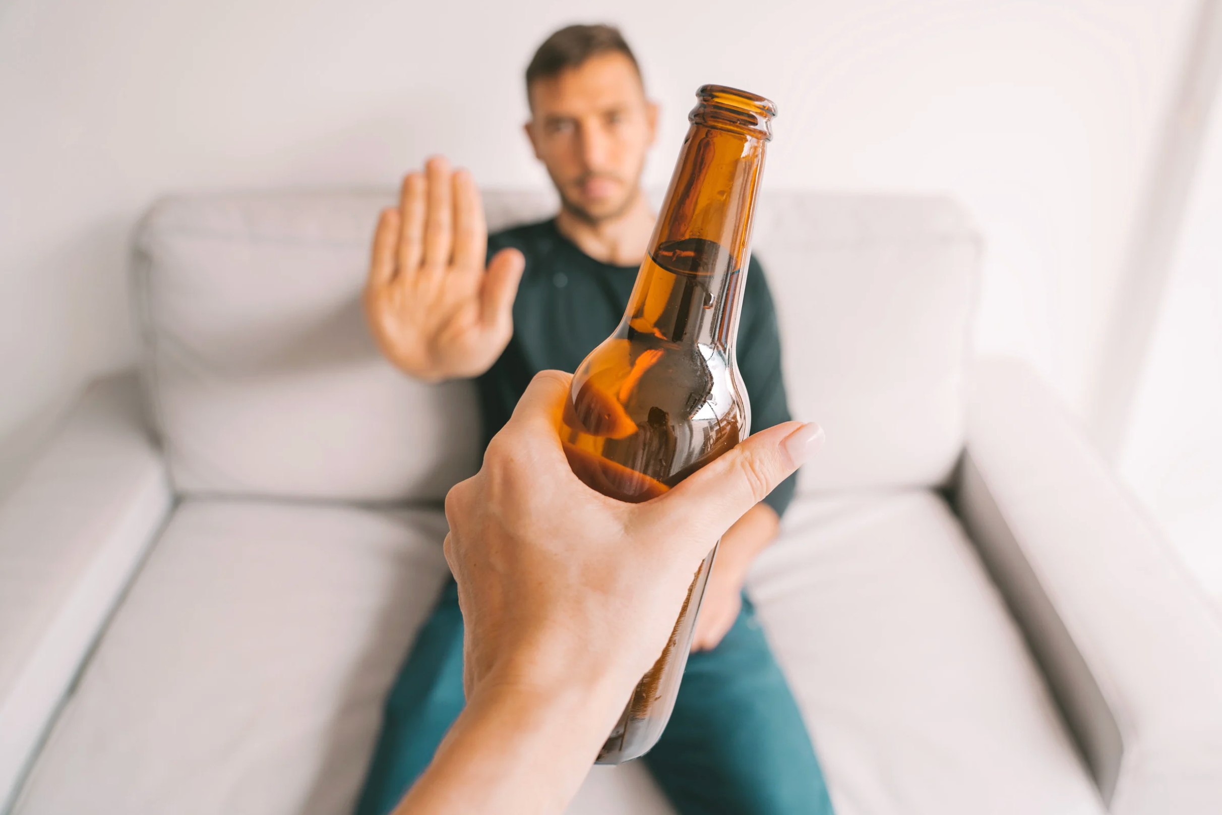 Young man sitting on a sofa declining a bottle of beer 