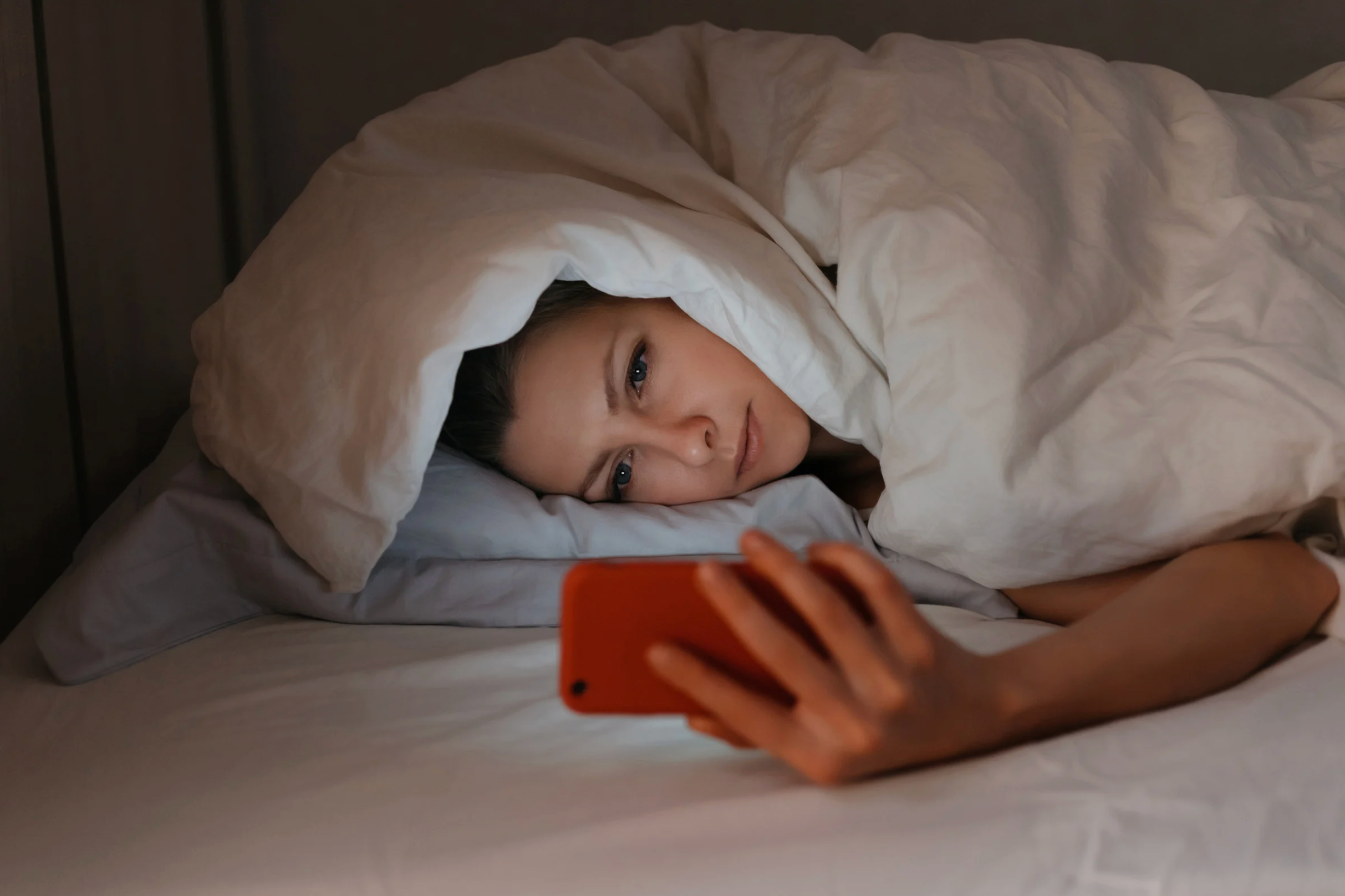 Young woman layin under a blanket in bed looking at her mobile phone