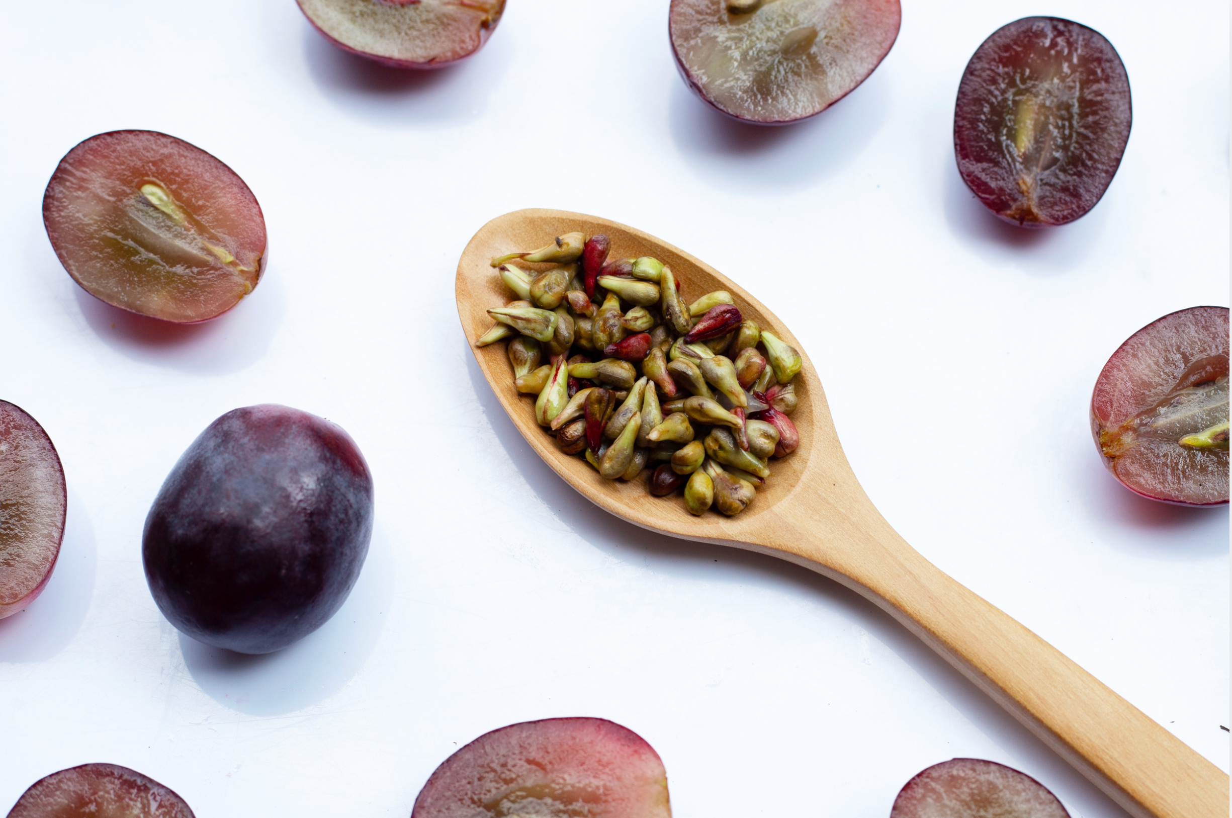 Wooden spoon with seeds surrounded by halved grapes on a white background
