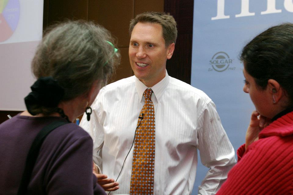 Dr. Murray in a white shirt and patterned tie standing between two people in an indoor setting with a Solgar sign in the background.