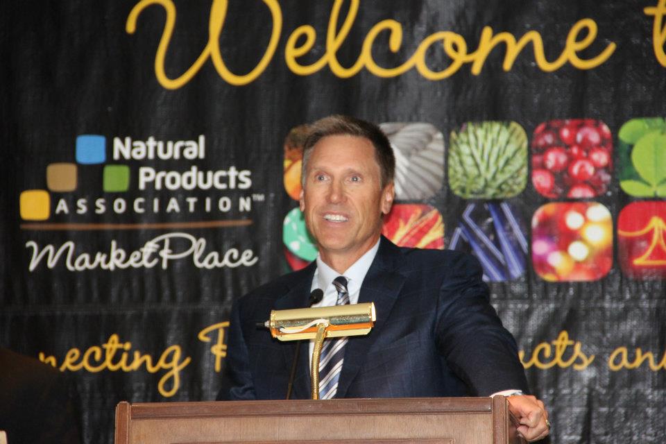 Dr. Murray speaking at a podium with a backdrop featuring the Natural Products Association logo and images of fruits and vegetables.