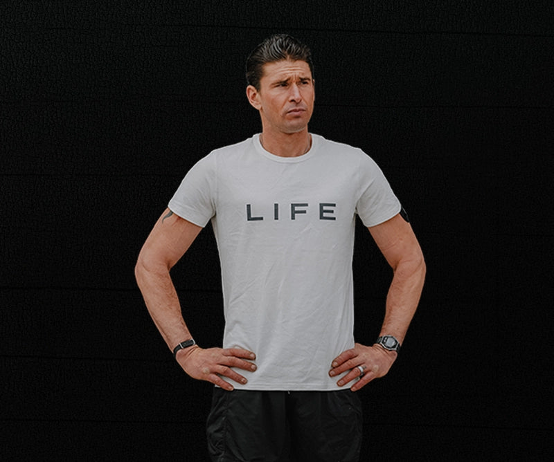 Ben Greenfield wearing a white t-shirt with the word 'LIFE' printed in black across the chest, posed against a dark background.