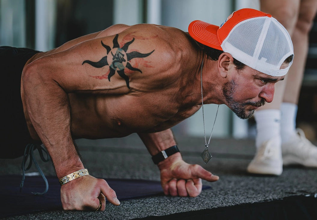 Ben Greenfield shirtless performing a push-up on a gym floor, wearing a red and white trucker hat backwards, a black tattoo on his upper back, and a silver necklace.